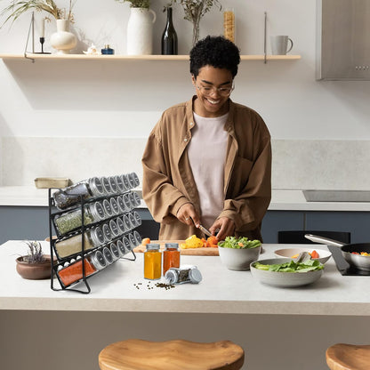4-tier spice rack organizer with 28 glass jars, labels, funnel, and marker on kitchen countertop, by The Willow Stone Collective.